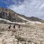 Three hikers with large backpacking packs on the Iceline Trail (part of the Yoho Valley Loop) in Yoho National Park. They are walking through a rocky landscape with glaciers on the mountain behind them.