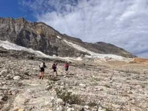 Three hikers with large backpacking packs on the Iceline Trail (part of the Yoho Valley Loop) in Yoho National Park. They are walking through a rocky landscape with glaciers on the mountain behind them.