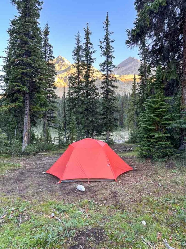 An orange tent at Little Yoho Camp in the Yoho National Park backcountry. You can see sun-dappled mountains in the background across a river. 