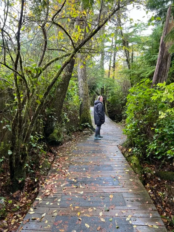 A woman in a black raincoat walks along a boardwalk through the forest on the way to Cox Bay Beach in Tofino.