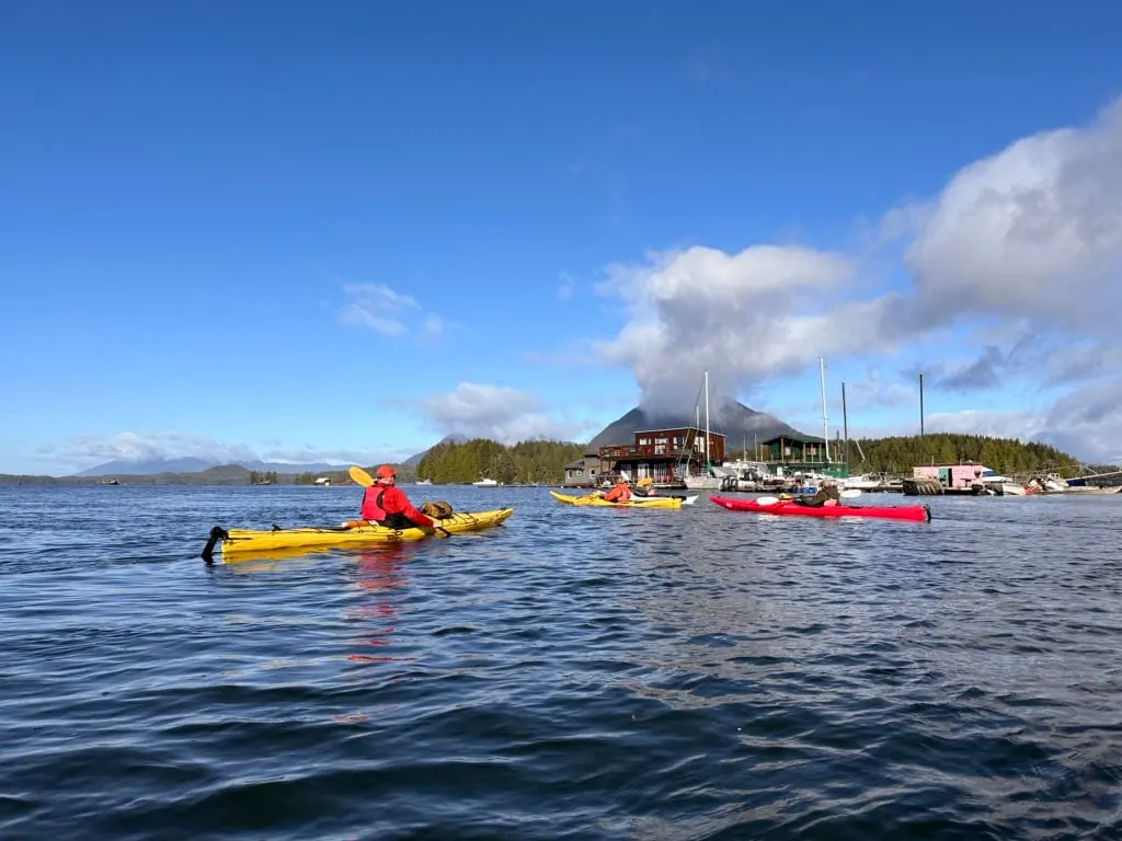 A group of kayaks paddlng past floating homes and moored boats in Tofino Harbour