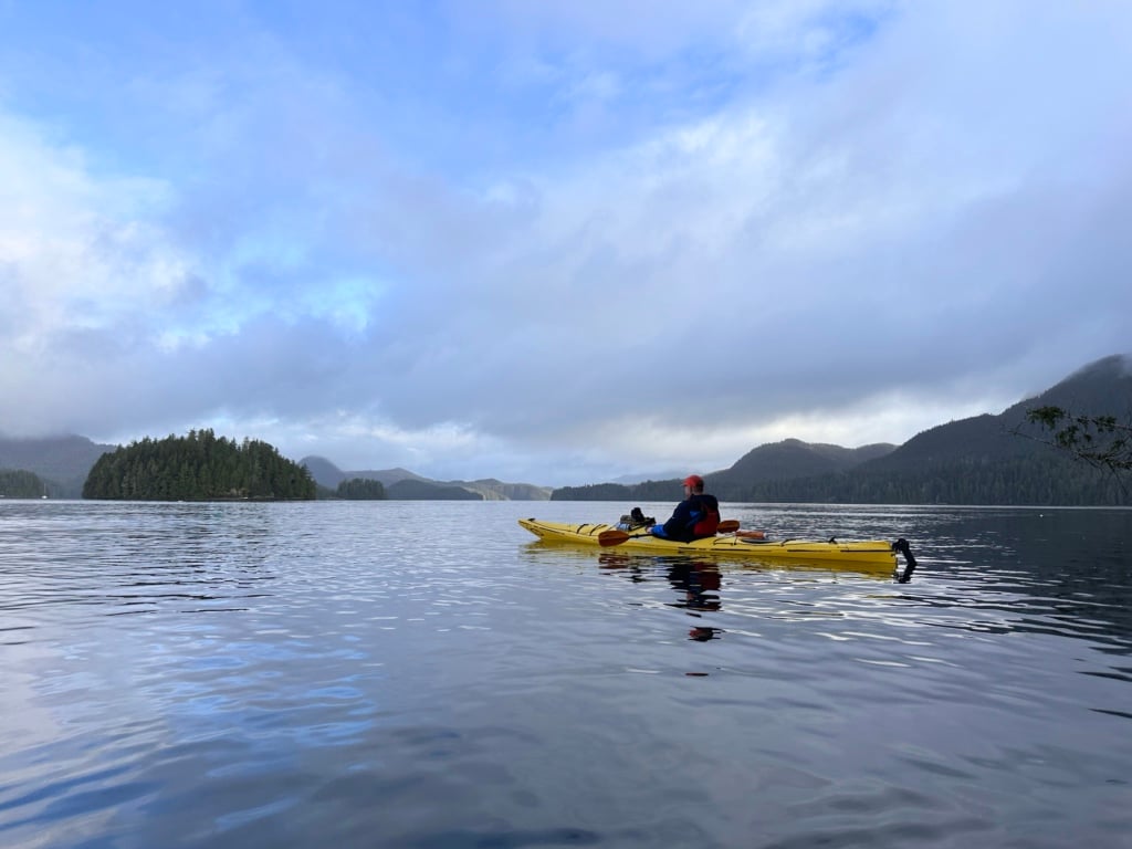 A man sits in a yellow kayak in Tofino Harbour with forested islands in the background. 