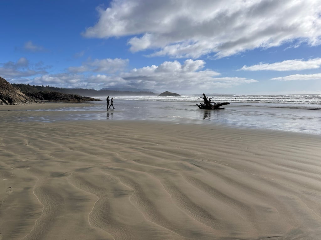 Two people walk along sand at Long Beach in Pacific Rim National Park. It is a sunny day. 