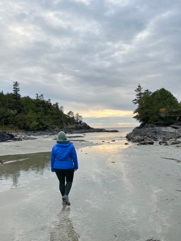 A woman wearing a rain coat walks along a beach in Tofino at sunset. 