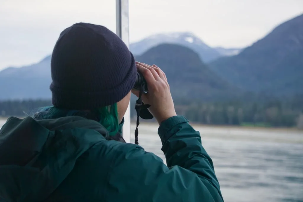 A woman looks through binoculars at a river and mountains