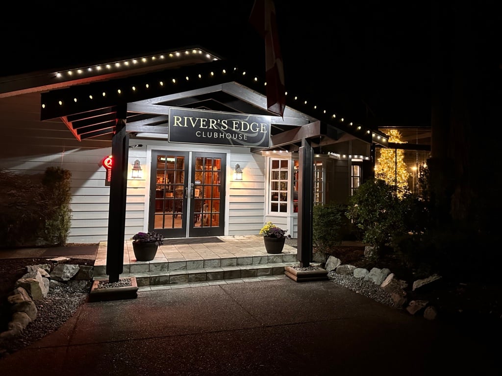 The exterior of the River's Edge Clubhouse Restaurant at the Sandpiper Resort at night. There are holiday lights along the roofline and a Christmas tree visible through the window. 