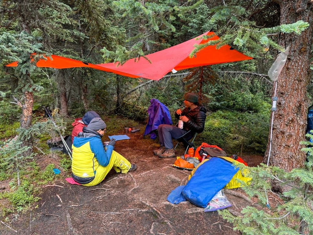A group of hikers eats under an orange tarp