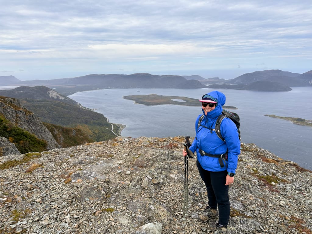A woman wearing a jacket with the hood up stands on top of Cape Blow Me Down near Corner Brook