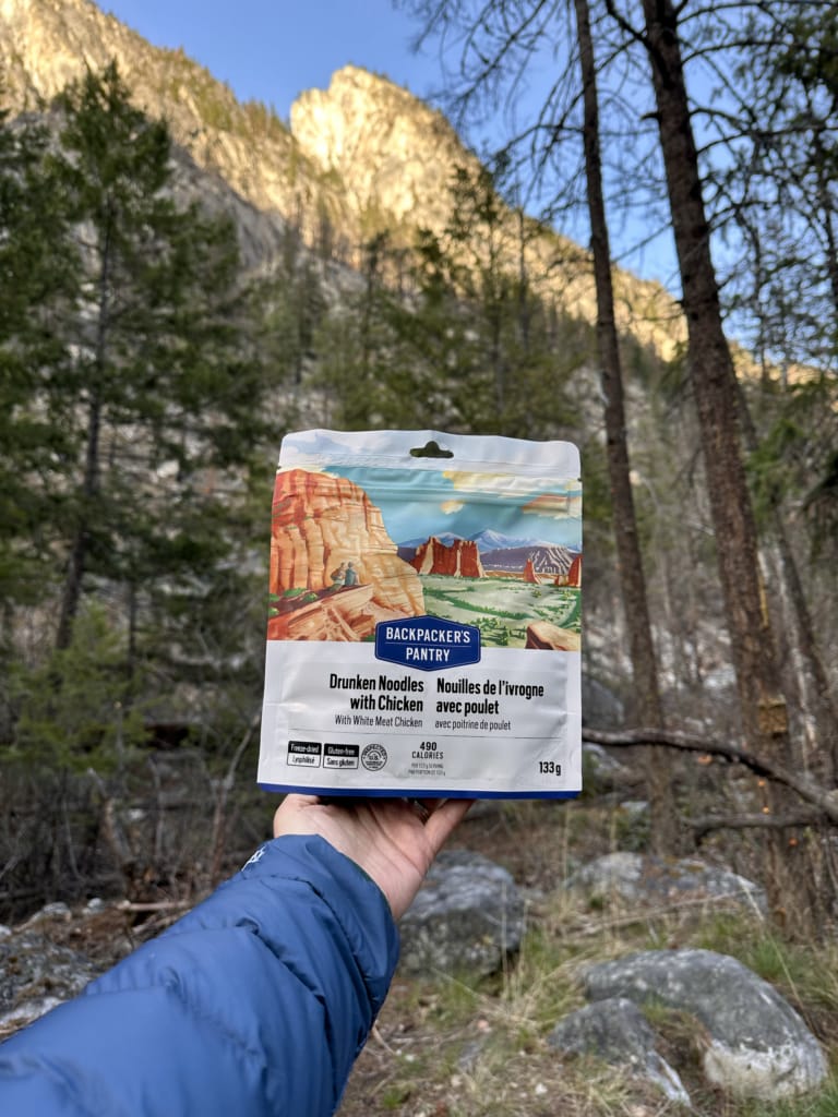 A package of Backpacker's Pantry Drunken Noodles with Chicken in front of a sunlit mountain