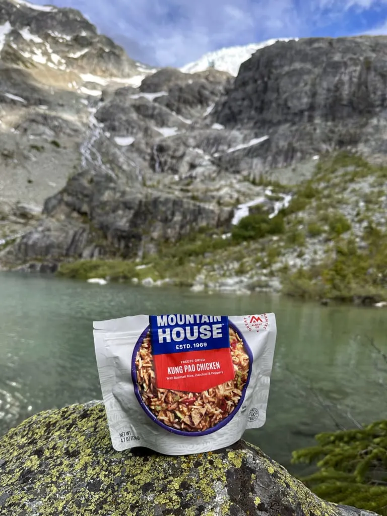 A package of Mountain House Kung Pao Chicken sits on a rock in front of an alpine lake
