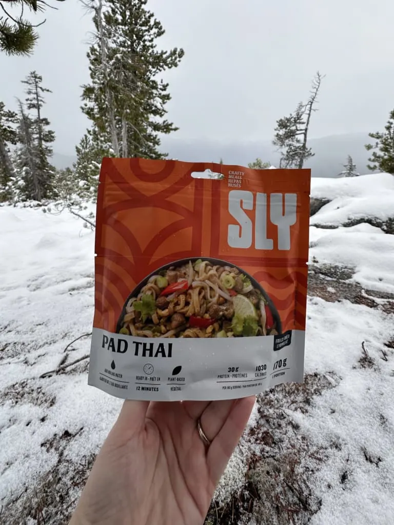 A hiker holds a package of SLY Pad Thai in front of a snowy trail
