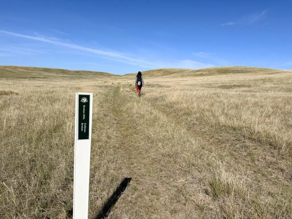 A woman walks past a trail marker in the grass on the Broken Hills Trail in Grasslands National Park