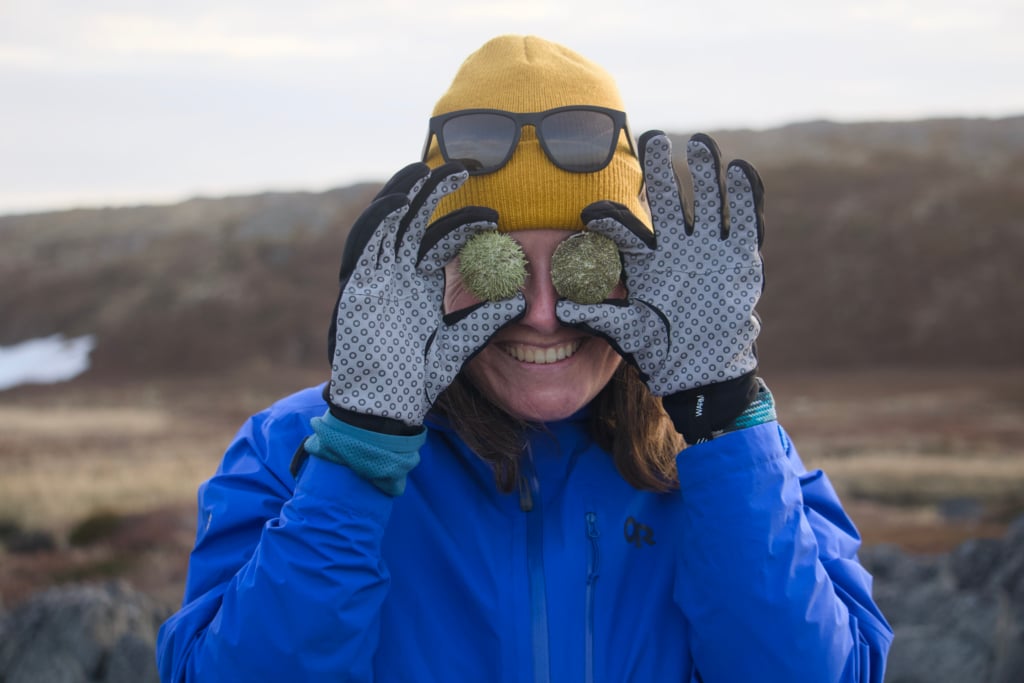 A woman wearing a yellow knit cap, gloves, and a blue jacket holds urchin shells over her eyes. 