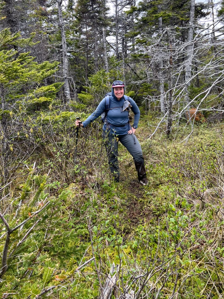 A hiker stands in a bog on the East Coast Trail. She has mud up to her knees. 